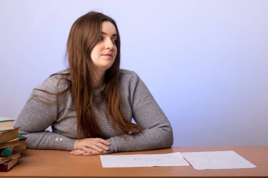 Genç bir öğrenci ya da öğretmen gri bir duvara karşı masada oturur ve yüzünü çevirir. Eğitim konsepti, koçluk. Sınav hazırlığı. Ev tatili. Avrupa görünümü.