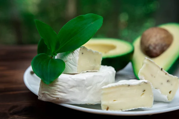 circle of sliced brie or camembert cheese with mold on a white plate. cut avocado on defocus background. Healthy diet. breakfast products. greenery nearby