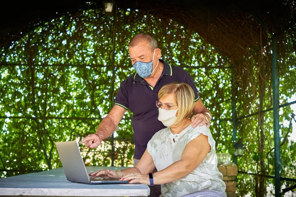 Senior couple browsing online with personal computer into a natural and green location during pandemic period using a protection face mask Stock Image