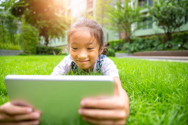 草の上に横たわって 公園でコンピューターのタブレットを見て小さなアジアの女の子 — ストック写真