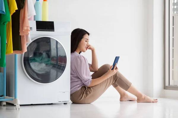 House wife sitting with cloth washing machine on floor watching smartphone during doing chore, Internet of thing concept