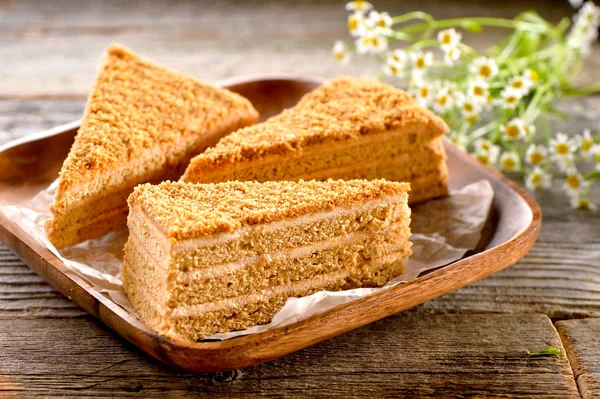 three pieces of honey cake with wooden platter,  baking  paper and field camomile flowers on textured wooden boards background