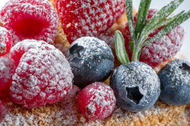 Taze ahududu, kızılcık, yabanmersini ve çilek üzerine krema serpiştirilmiş şeker. Meyveli kekin üstüne yakın plan biberiye yaprakları beyaz arka plana serpiştirilmiş. 