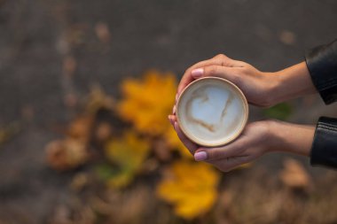 Bir kızın ellerinde bir fincan kahve var. Üst manzara, sadece siyah ceketin kollarına sahip kadın avuçları görülebilir. Arka planda biberiyede sonbahar yaprakları olan bir zemin var..