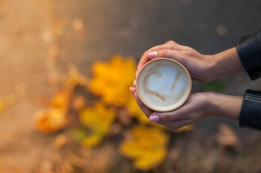 Bir kızın ellerinde bir fincan kahve var. Üst manzara, sadece siyah ceketin kollarına sahip kadın avuçları görülebilir. Arka planda biberiyede sonbahar yaprakları olan bir zemin var..
