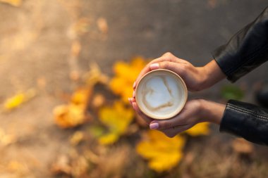 Bir kızın ellerinde bir fincan kahve var. Üst manzara, sadece siyah ceketin kollarına sahip kadın avuçları görülebilir. Arka planda biberiyede sonbahar yaprakları olan bir zemin var..