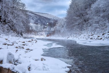 Kışın dağlarda, karla kaplı Noel ağaçları kıyılar boyunca buzla kaplı bir nehir ve mavi gökyüzünde bulutlar..