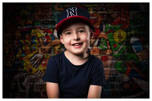 Крупный план портрет маленького мальчика в повседневной одежде, кепка, глядя улыбаясь прямо в камеру, очаровательный ребенок стоит напротив графитти стены. — стоковое фото