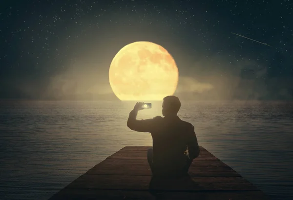 A man sitting on a pier looks at the moon and photographs it on