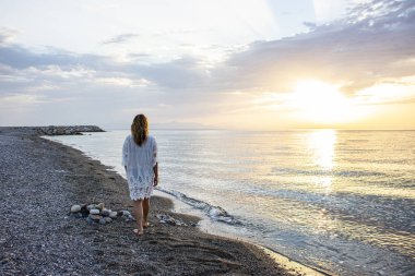 Beyaz elbiseli bir kız güneş doğarken sahilde yürüyor..