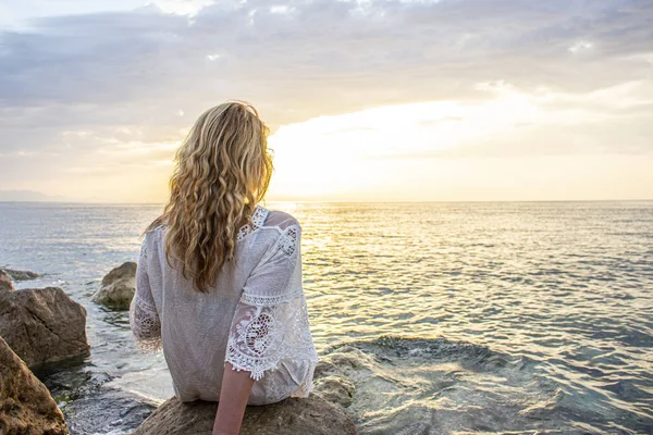 Altın saçlı bir kız denizde bir taşın üzerinde tek başına oturur ve güneşin doğuşunun tadını çıkarır..