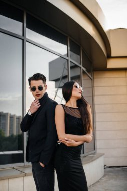 Siyah elbiseli ve gözlüklü güzel, şık bir çift genç gün batımında bir ofis binasının arka planında duruyor. Moda ve tarz