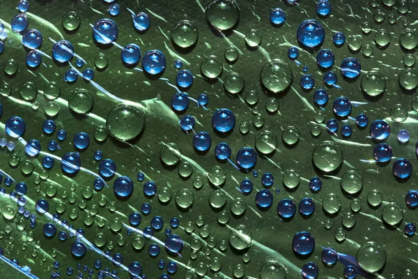 Transparent green and blue drops of water flow down on the glass  surface of a green background. Close-up.