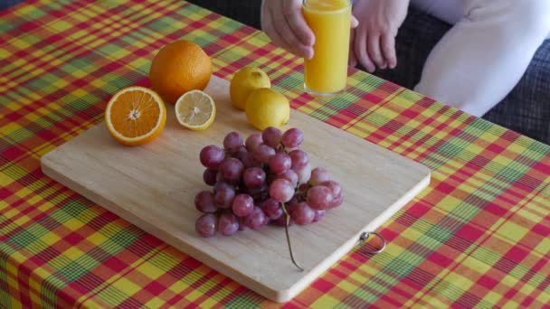 白いパンツを着た無名の男が座ってオレンジジュースのグラスをテーブルの上に置いたこの健康的な食事は マドラスパターンテーブルクロス 木製のプレート 生の新鮮な果物で構成されています — ストック動画
