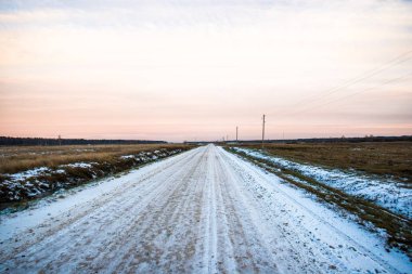 Gün batımında tarlalardan karla kaplı toprak bir köy yolu. Arka planda orman. Letonya