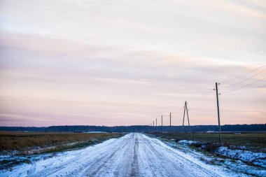 Gün batımında tarlalardan karla kaplı toprak bir köy yolu. Arka planda orman. Letonya