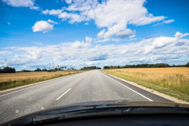 Mavi gökyüzüne karşı kırsal alanlarda bir asfalt yol, arabanın penceresinden bir görünüm. Estonya