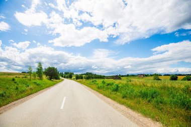 Güneşli bir yaz gününde arka planda bir orman ile yeşil ülke alanları ile bir asfalt yol, Letonya