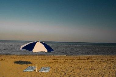 Bir çift şezlong ve kumsal şemsiyesi mükemmel ıssız bir plajda, arkasında mavi deniz ve gökyüzü olan.