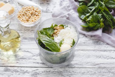 Beyaz ahşap masada peynir, çam fıstığı, zeytinyağı, fesleğen, sarımsak bir blender pesto sos yapma süreci.