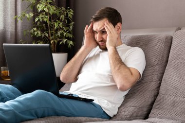 Stresli Kafkasyalı, stresli, ev ofisinden çalışan, bilgisayar ve internet kullanarak koruyucu maske takan bir adam. Uzaktan çalışma, ofis işyerinde kanepe üzerinde serbest çalışma. Coronavirus covid-19 karantinası