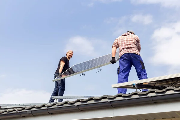 Mutlu elektrik mühendisleri mavi gökyüzüne karşı güneş panelini çatıda tutuyor. Alternatif enerji teknolojisinin geliştirilmesi. Ekolojik kavram.