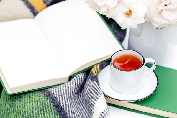 Vintage cup of tea near open book, flowers white wild rose in vase, warm plaid on white table outside in garden. Romantic leisure breakfast with nature background. Reading in provence cafe.