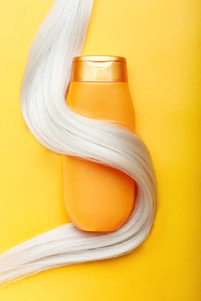 Shampoo bottle wrapped in lock of blonde hair on orange color background. Gold bottle shampoo in dyed hair strand. Hair care cosmetics bath beauty products hair treatment. Top view.