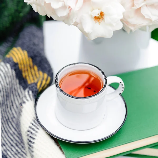 Cup of tea on books, flowers white wild rose in vase , warm plaid on white table outside in garden. Romantic leisure breakfast with nature background. Cozy home backyard or provence cafe.