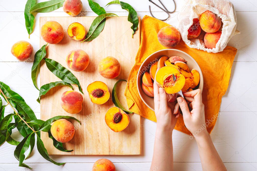 Female hands cutting fresh sweet peaches. Peaches whole fruits leaves, half peach, peach slices on white wooden kitchen table. Recipe making peach jam, cooking peach dessert on cutting board.Flat lay.