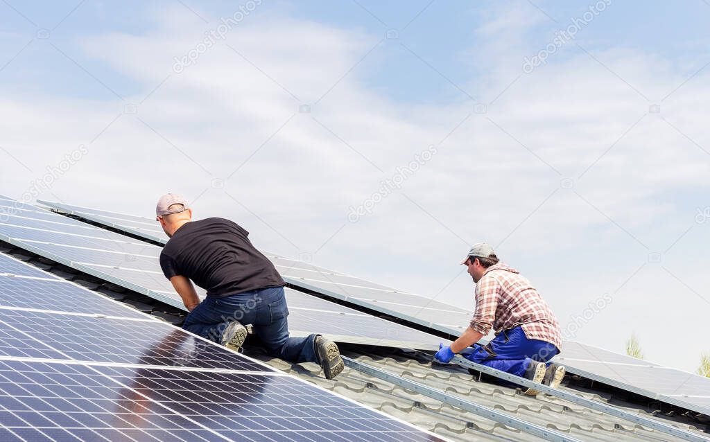 Process of building a solar station. Electrical engineers mans are working installing solar panels on solar station on house roof against blue sky. Alternative energy ecological concept