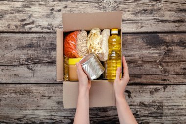 Kadın eller bağış kutusunu paketliyor. Tahta masanın üzerinde zımba ürünleri var. Bağış yapan kadın yiyecek kutusu alıyor. Yemek dağıtım konsepti. Bağışlar markette konserve yiyecek. Üst görünüm