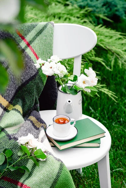 Summer breakfast in garden. Cup of tea on books, flowers white wild rose in vase teapot, warm plaid on white chair outside in garden. Romantic provence leisure breakfast with nature background.
