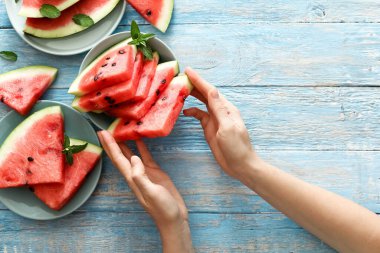 Fotokopi alanı olan mavi bir ahşap arka planda dişi ellerinde taze kırmızı karpuz dilimi. Arka planda karpuz dilimi ve nane şekeri. Üst görünüm.