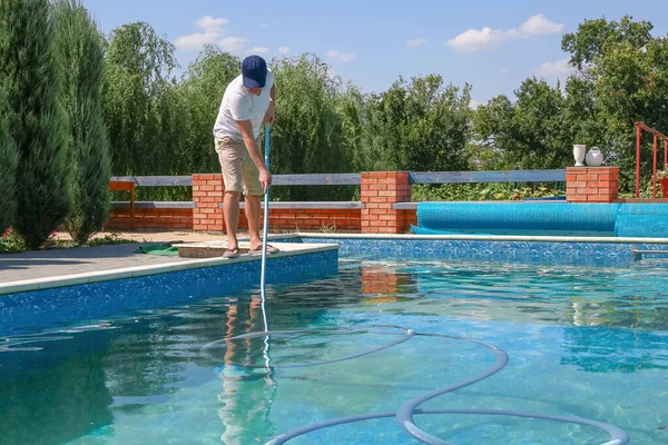 Cleaner of swimming pool. Man cleaning outdoor swimming pool with vacuum tube cleaner in summer. Seasonal preparations. Cleaning systems for swimming pools. Dirty outdoor pool.