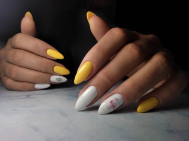 Beyaz tasarım ve çizimler ile uzun sivri çivi sarı jel cila. Beyaz tasarımlı manikür ve beyaz arka plan üzerinde pembe flamingo ve yeşil dal çizilmiş.