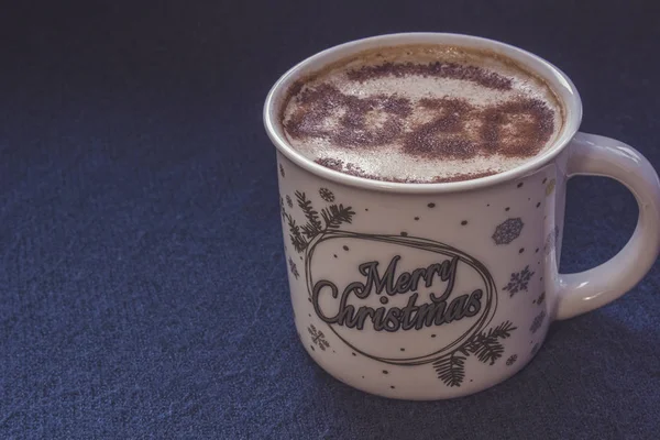 Mug of cappuccino with cinnamon on a dark blue background.Cinnamon powder was sprinkled with 2020 figures on coffee foam.Symbol of New year in a cup on coffee foam on woolen fabric.