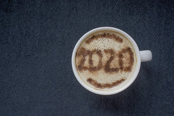 Mug of cappuccino with cinnamon under a cyanide color on a dark background.Cinnamon powder was sprinkled with 2020 figures on coffee foam.Symbol of New year in a cup on coffee foam on woolen fabric.