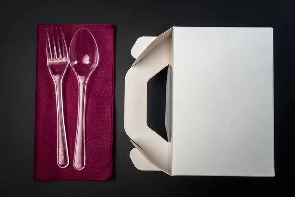 White Cardboard Fast Food Box, Packaging For Lunch and plastic fork and spoon on purple serviette. On black Background Isolated. Ready For Your business design