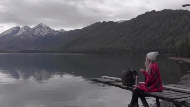 Junge Frau sitzt auf einem alten Steg am Ufer des Bergsees und trinkt — Stockvideo