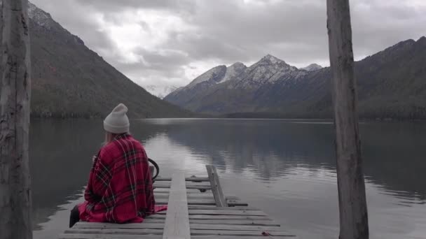 Ung kvinna sitter på en gammal brygga på stranden av fjällsjö och drycker — Stockvideo