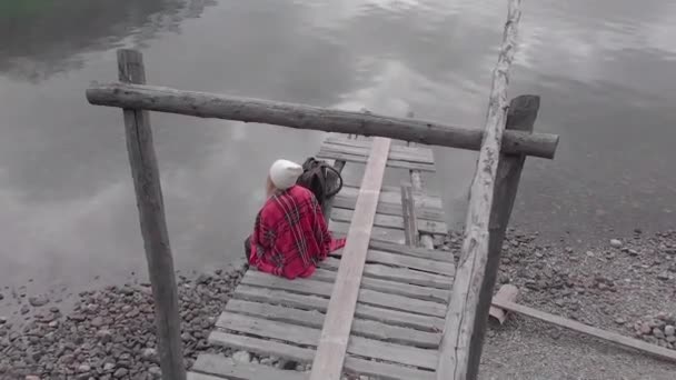 Giovane donna siede su un vecchio molo sulla riva del lago di montagna e beve — Video Stock