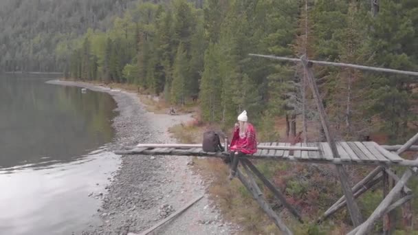 Jeune femme assise sur une vieille jetée au bord du lac de montagne et boissons — Video