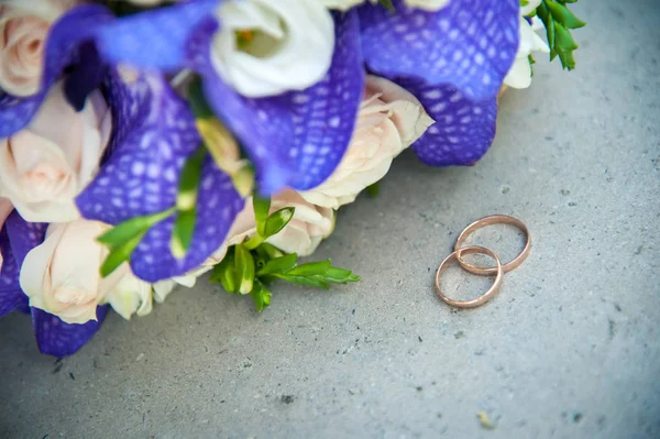 two gold wedding rings lie side by side on a concrete background and next to them in the corner is visible part of the bride\'s wedding bouquet of peach roses and large purple flowers with spotted petals