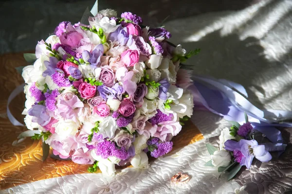 Wedding bouquet of the bride tied with ribbons of white, pink, lilac and purple flowers, roses and herbs, boutonniere groom and two gold wedding rings on the blanket