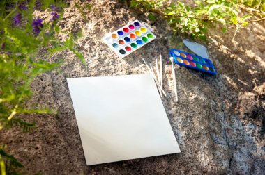 watercolor paints and a few brushes lie on a large stone, next to a clean sheet of white paper for drawing clipart
