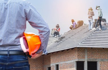 çatı evi konut geliştirme konsepti ise çatıya beton fayans yükleme işçileri ile şantiye altında yeni ev binasında sarı güvenlik kask tutan yerleşik mühendis