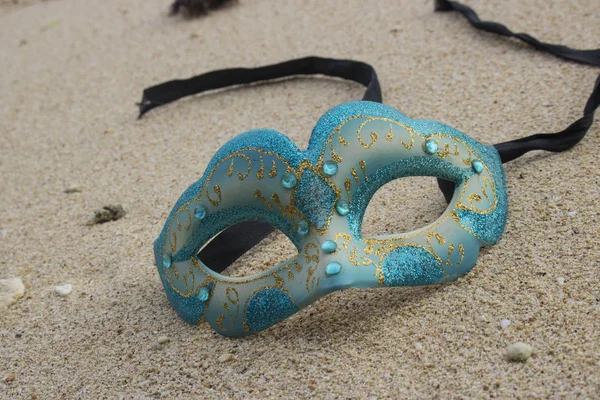 blue masquerade mask lying on sand, beach party concept