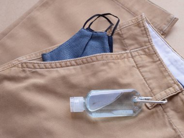 Askılı alkol şişesi jeli ve cep kahverengi uzun pantolon arka planında mavi kumaş maske. Evden her çıkışında kirliliği ve virüsleri önlemek için kullanılmalıdır. Sağlık hizmetleri, yeni normal kavramlar