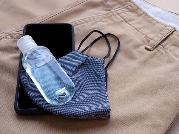 Alkol şişesi jeli ve kahverengi uzun pantolon arka planında akıllı telefonlu mavi kumaş maskeli. Evden her çıkışında kirliliği ve virüsleri önlemek için kullanılmalıdır. Sağlık hizmetleri, yeni normal kavramlar